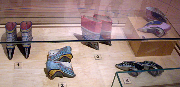 [Five different pairs of colorful shoes all with heels in a glass case. Two pair are upright. Two pair are laying on their sides. And one pair has one shoe upright and one on its side.]